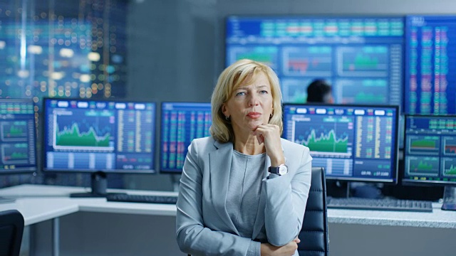 股票市场领先分析师努力思考解决财务问题。在她身后的人工作和显示器显示图表和数字。视频素材
