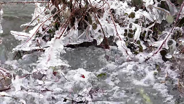 一条美丽清澈的小河从积雪中流淌而出，声声融解。流动的水形成了有趣的形状，因为它在原始的冰晶，4k冬季背景视频素材