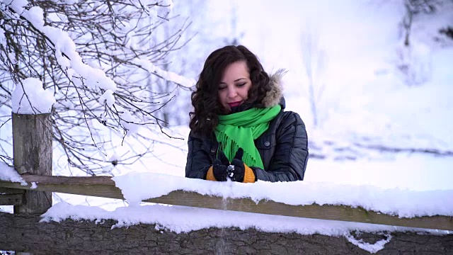 年轻美丽的女孩在白雪覆盖的公园里。她站在木栅栏附近。女孩把雪捧在手里，朝他吹了吹。高高兴兴地把雪堆起来。视频素材