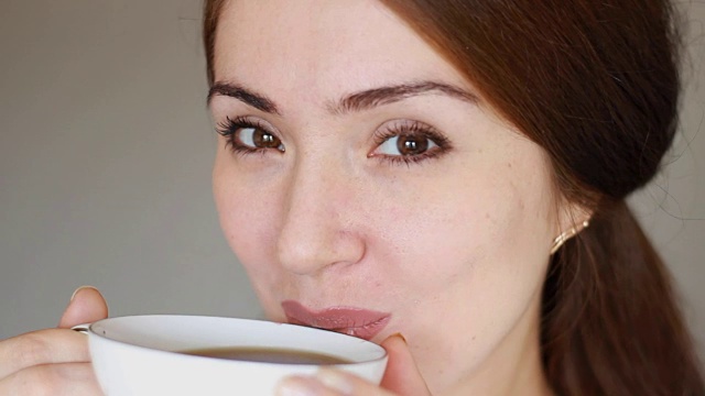 年轻漂亮的女人从一个白色的杯子里喝了一杯热饮，看着镜头。女孩享受着香味，抿了一口。特写镜头视频素材