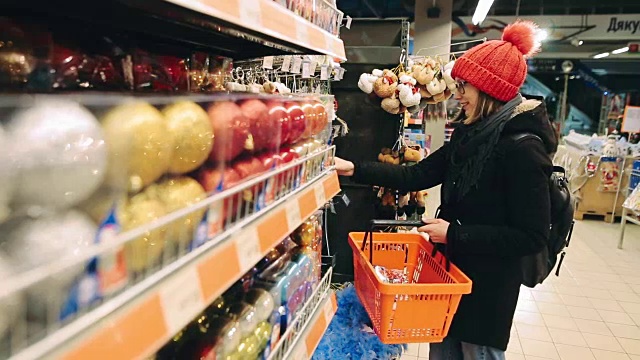 漂亮的年轻女孩戴着眼镜选择圣诞装饰装饰圣诞树。买主在商店里找到一个便宜的玩具，把它们放在购物篮里。“黑色星期五”的概念视频素材