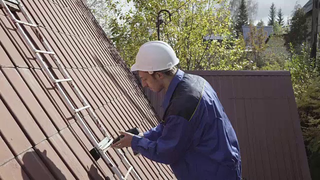 秋天，一名戴着白色建筑头盔、穿着蓝色工作服的建筑工人在一所乡村住宅的屋顶上安装了一架梯子，他使用了一个4k无绳螺丝刀。视频素材