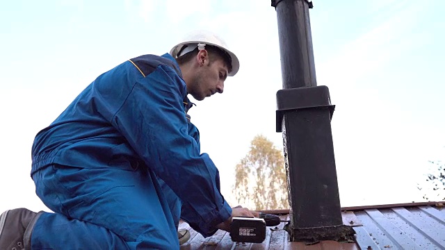 一个年轻的工人在蓝色工作服和一个建筑头盔修理烟囱与无绳钻近距离4k。视频素材