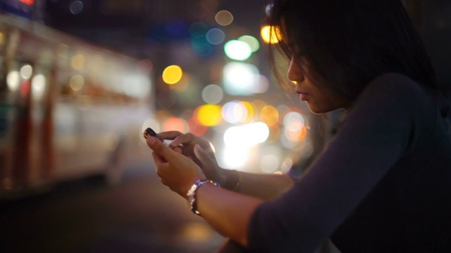 亚洲女孩在公交车站附近的夜间街道上使用智能手机视频素材