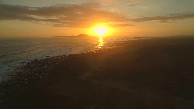 空中镜头:当浪花到达岩石海岸时，夕阳正落在火山后面。视频素材