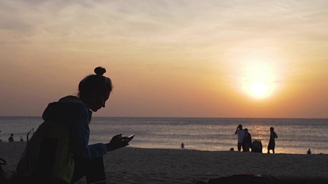 夕阳西下时，一个悲伤的女孩坐在海滩上写着电话短信。视频素材