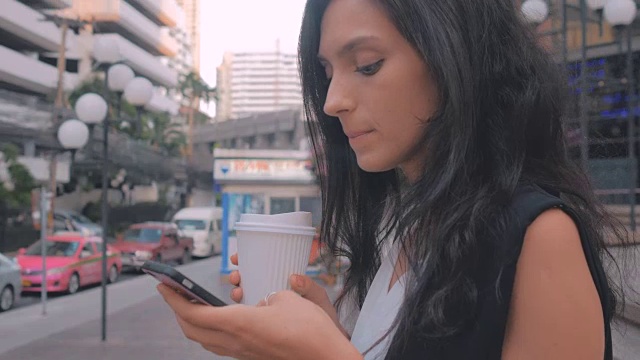 拿着智能手机喝着咖啡在城市里散步的女人，用稳定器拍摄视频素材