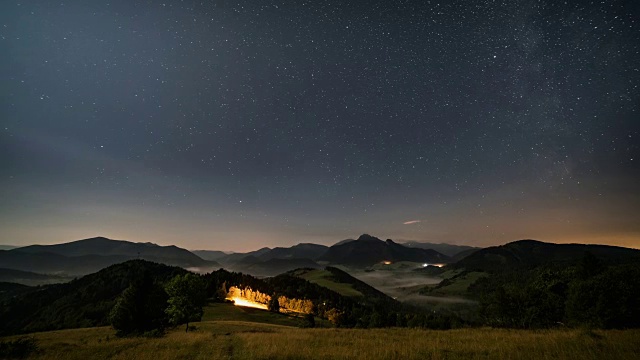 在月光下，星星在山峦和朦胧的乡村景色的夜空中移动。从黑夜到白天的时间流逝摄影车拍摄视频素材