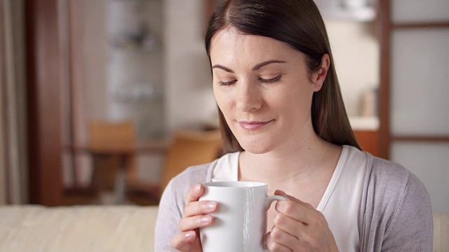 年轻女子在家里的沙发上休息。漂亮的女人在客厅用杯子喝咖啡或茶视频素材