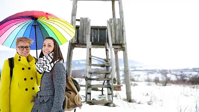 两个快乐的女人背着背包，撑着雨伞，走在冬日的雪地里视频素材