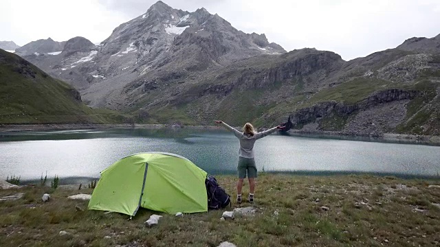 无人机拍摄，一名年轻女子在徒步旅行站在她的帐篷附近美丽的山湖双臂伸展视频素材