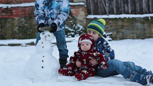 可爱的小男孩和他的两个哥哥，坐在户外的雪地里，雪人在他们旁边视频素材