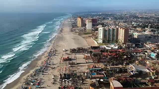 航拍向后飞行在繁忙的海滩，汹涌的海浪，和度假村在罗萨里托，墨西哥。视频素材