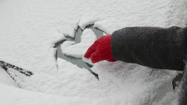 一辆被雪覆盖的汽车上的爱情告白——一个女人正在一辆被雪覆盖的汽车的挡风玻璃上画一个心形视频素材