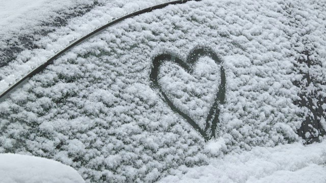 一辆被雪覆盖的汽车上的爱情告白——一个女人正在一辆被雪覆盖的汽车的侧窗上画一个心形视频素材