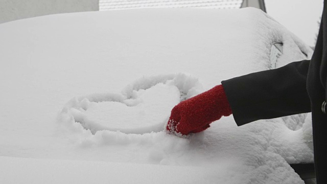 一辆被雪覆盖的汽车上的爱情告白——一个女人正在一辆被雪覆盖的汽车的挡风玻璃上画一个心形视频素材