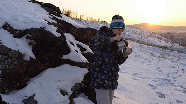 在城市附近的小山上，一位年轻女子从保温瓶里喝茶或喝咖啡。视频素材