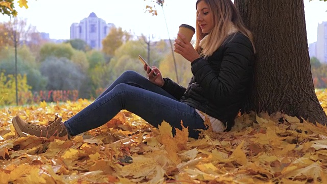 坐在枯黄落叶旁的Pan Woman，使用应用程序和喝咖啡视频素材