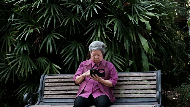 亚洲老妇人拿着手机在花园里。老年女性在公园里用智能手机上的应用发短信。老年人用手机连接社交网络视频素材