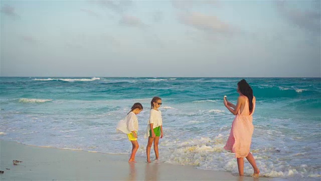 在温暖的夜晚，可爱的小女孩和年轻的妈妈在热带海滩上。妈妈在日落上给漂亮的孩子们拍了一张照片视频素材