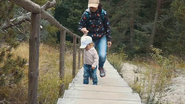 一家游客妈妈和儿子爬上台阶，在树林里露营。年轻的妈妈抱着她的儿子，帮助婴儿爬上楼梯的台阶。家人一起旅行，户外踏踏大自然。视频素材