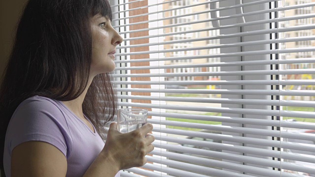 一位年轻女子正站在窗边喝着干净的淡水。女孩喝了一杯水，看着窗外。黑发的女人。视频素材