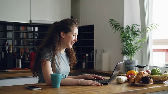 Curly相当积极的白人女人坐在桌子上用笔记本电脑上skype，她正在谈论坐在厨房靠近窗户，一杯咖啡站在笔记本电脑附近视频素材