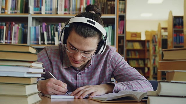 年轻的白人男学生戴着大耳机坐在图书馆的桌子旁。他周围有很多书，他在微笑着听音乐和写作视频素材