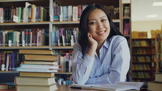 年轻美丽的亚洲女学生的肖像坐在桌子与图书馆的教科书看着相机。她满脸堆笑。视频素材