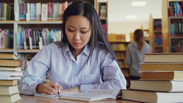 年轻美丽的亚洲女学生坐在桌子周围的书籍在大图书馆重写课本准备考试视频素材