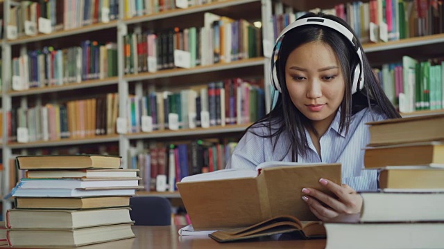 相当积极的亚洲青少年学生戴着耳机听音乐坐在大图书馆的桌子上拿着书和写总结视频素材