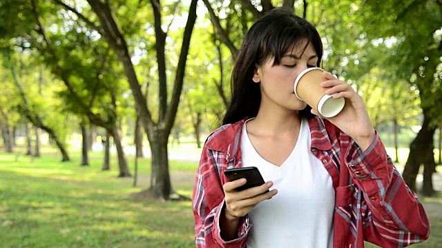 美丽的女人喝咖啡和打字在她的智能手机在公园里，有吸引力的女性使用她的触摸屏手机发送按摩视频素材