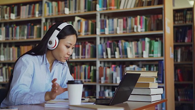 美丽的亚洲女学生听音乐耳机坐在书桌在图书馆工作的学校项目。年轻女孩在笔记本电脑上打字，喝咖啡，在抄写本上写字视频素材