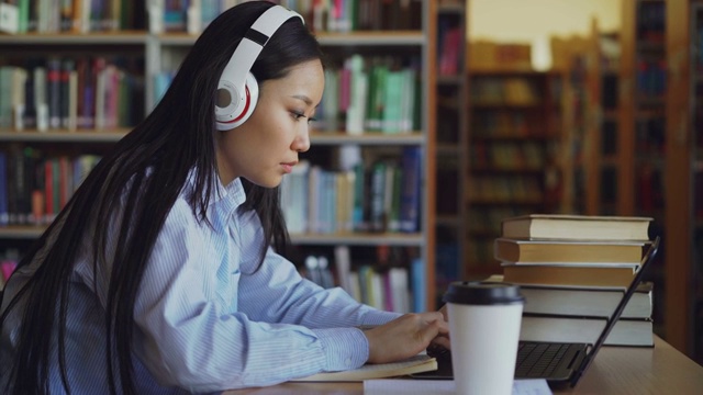 美丽的亚洲女学生戴着耳机坐在图书馆的书桌前专注于学校项目。年轻女孩在笔记本电脑上打字，喝咖啡，在抄写本上写字视频素材