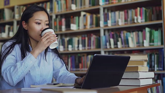 年轻的亚洲学生女孩坐在大学图书馆的一堆书的桌子上，在笔记本电脑上工作，喝咖啡微笑着看着别处，准备考试视频素材