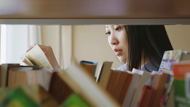 迷人的亚洲学生女孩站在书架旁边的书在大学图书馆拿书翻页阅读它视频素材