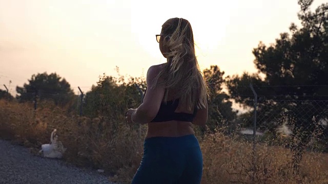 日出时，跟随在乡村小路上慢跑的运动女孩。年轻的女人戴着太阳镜在户外跑步，听着智能手机里的音乐。健康积极的生活方式。慢镜头后景特写视频素材