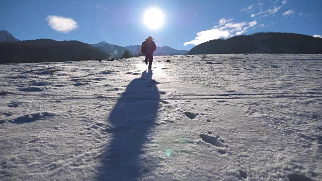 在阳光明媚的冬日，一个面目全非的家伙在雪地上慢跑。年轻人在白雪覆盖的草地上奔跑。背景是阳光和蓝天。假日或假期的概念。后视图慢动作视频素材