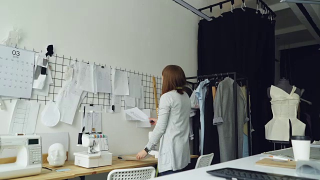 女服装设计师从工作室的桌子上取草图，并把它们与其他图纸的女性服装放在墙上。创造性思维的概念。视频素材