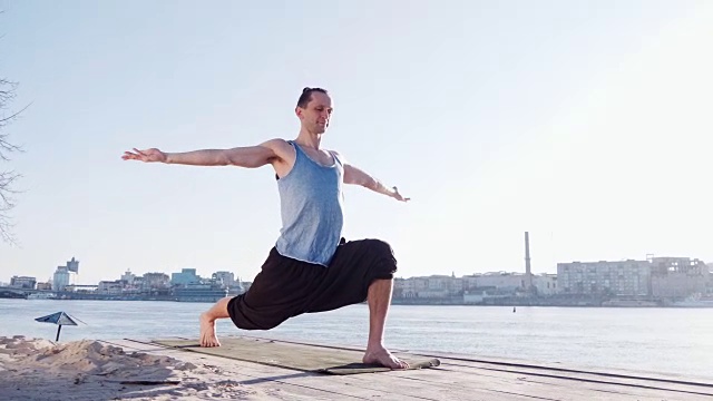 年轻的白人男子放松练习瑜伽健身运动在海滩附近平静的河流与城市的背景视频素材