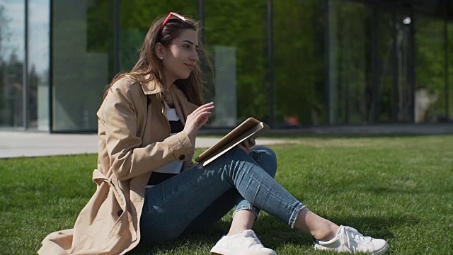 年轻的女学生坐在户外的草地上微笑着使用平板电脑。视频素材