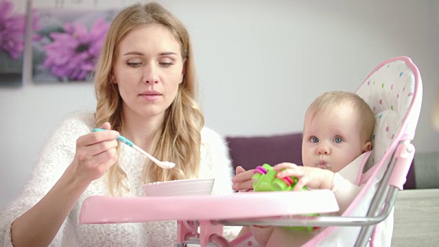 拿着玩具吃婴儿碗里的食物泥的孩子。坐在椅子上喂婴儿的妇女视频素材