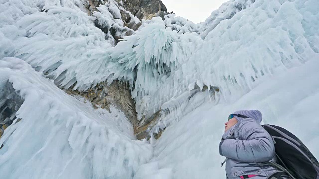 女人在贝加尔湖冰面上的旅行。冬岛之旅。女孩在冰岩石下行走。游客看着美丽的冰洞。极限跋涉和步行。背包客在大自然中休憩。视频素材