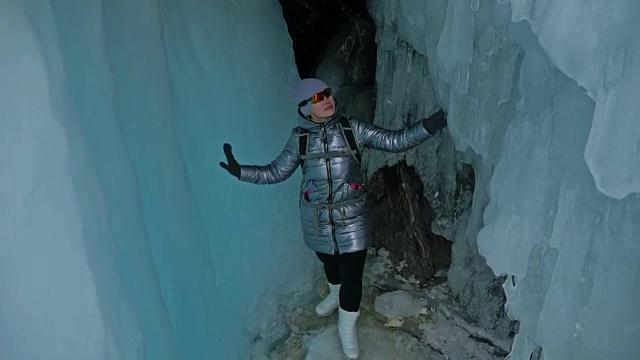 贝加尔湖冰洞上的女游客。冬岛之旅。女孩背包客正在冰洞中行走。游客看着美丽的冰洞。徒步旅行者穿银色夹克，背包，led灯。视频素材