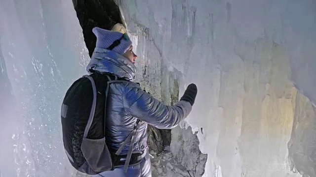 贝加尔湖冰洞上的女游客。冬岛之旅。女孩背包客正在冰洞中行走。游客看着美丽的冰洞。徒步旅行者穿银色夹克，背包，led灯。视频素材