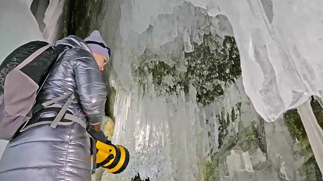 贝加尔湖冰洞上的女游客。冬岛之旅。女孩背包客正在冰洞中行走。游客看着美丽的冰洞。徒步旅行者穿银色夹克，背包，led灯。视频素材