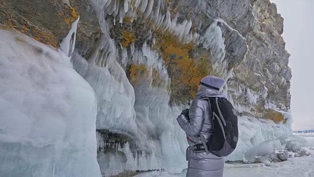 女人在贝加尔湖冰面上的旅行。冬岛之旅。女孩在冰岩石下行走。游客看着美丽的冰洞。极限跋涉和步行。背包客在大自然中休憩。视频素材