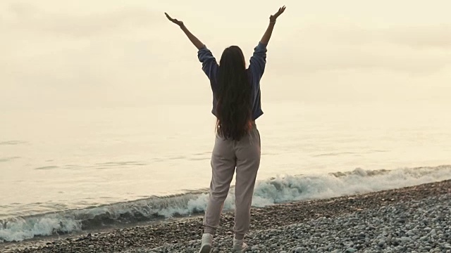 迷人的黑发女子举起双手，站在卵石滩上，夕阳西下视频素材