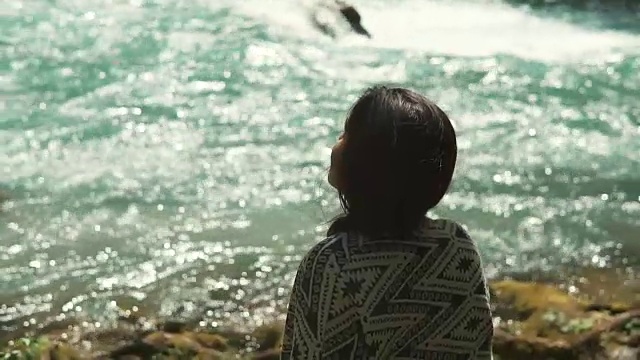 在阳光明媚的日子里，沉思的女孩坐在河边，从她的背后看视频素材