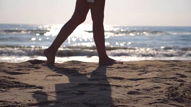 在阳光明媚的日子里，女性的脚在海滩上行走，海浪在背后。年轻女子的双腿踩在沙滩上。暑假或假日概念。侧视图慢动作关闭低角度视图视频素材